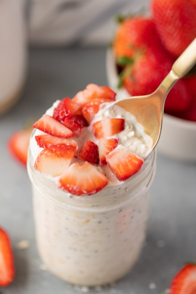 A mason jar of strawberry overnight oats with a gold spoon taking a bite out of it