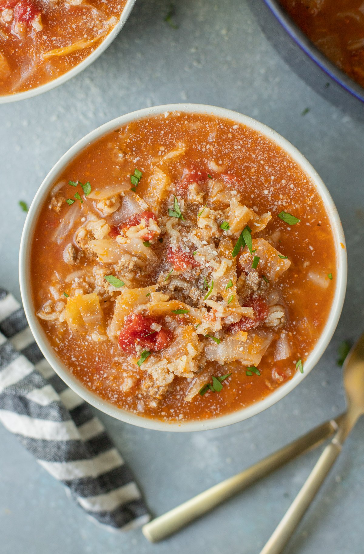 A bowl of hamburger cabbage soup with another bowl of soup and spoons on the side on a grey counter