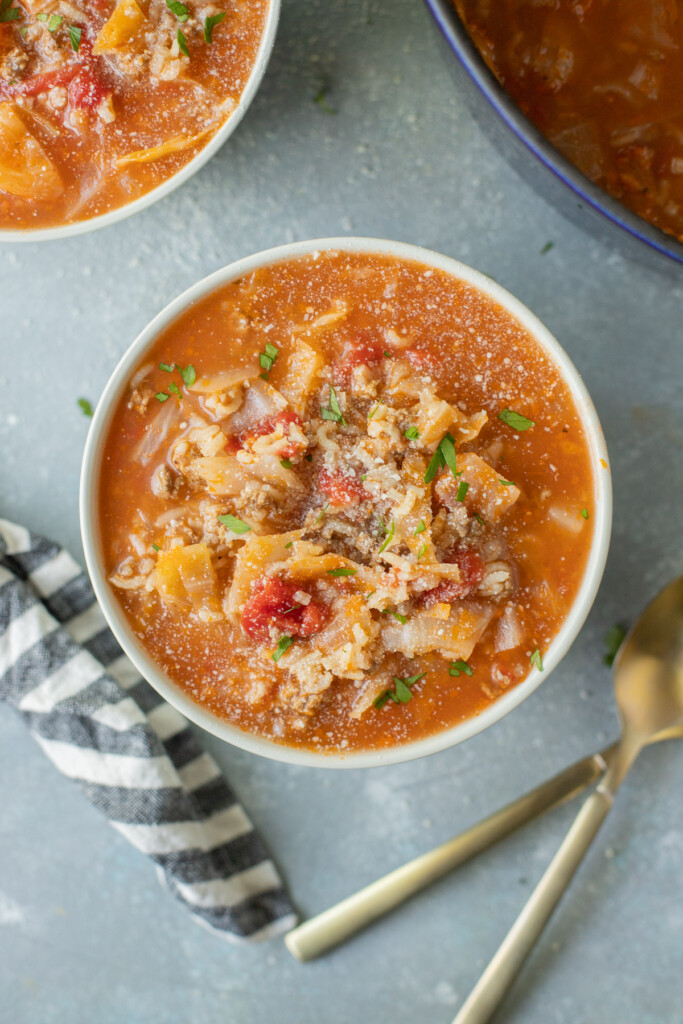 A bowl of stuffed cabbage soup with another bowl of soup and spoons on the side on a grey counter