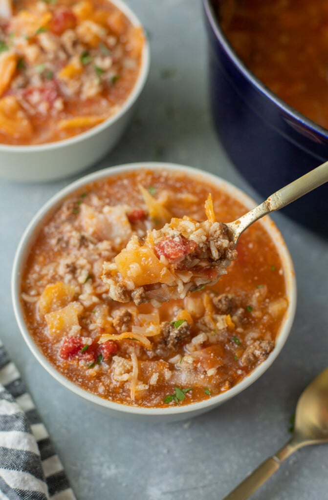 A spoon taking a bite out of a bowl of cabbage roll soup with another bowl and dutch oven of the soup in the background on a blue counter
