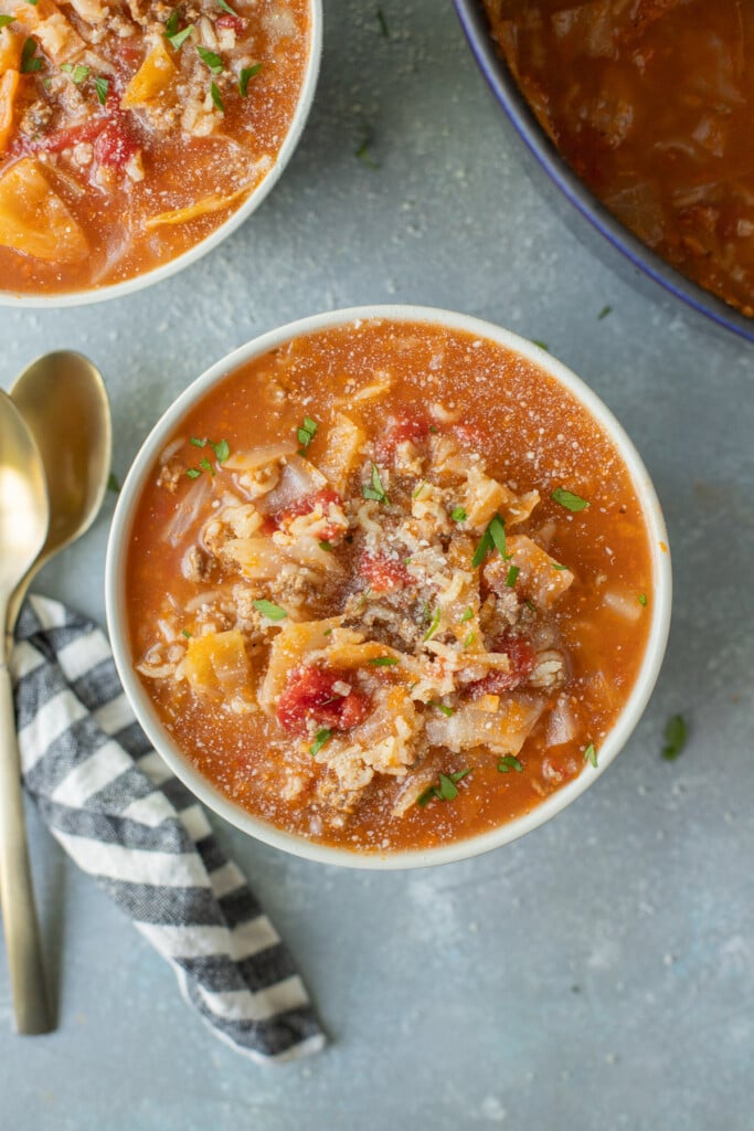 A bowl of stuffed cabbage soup with another bowl of soup, a dutch oven, and spoons on the side on a grey counter