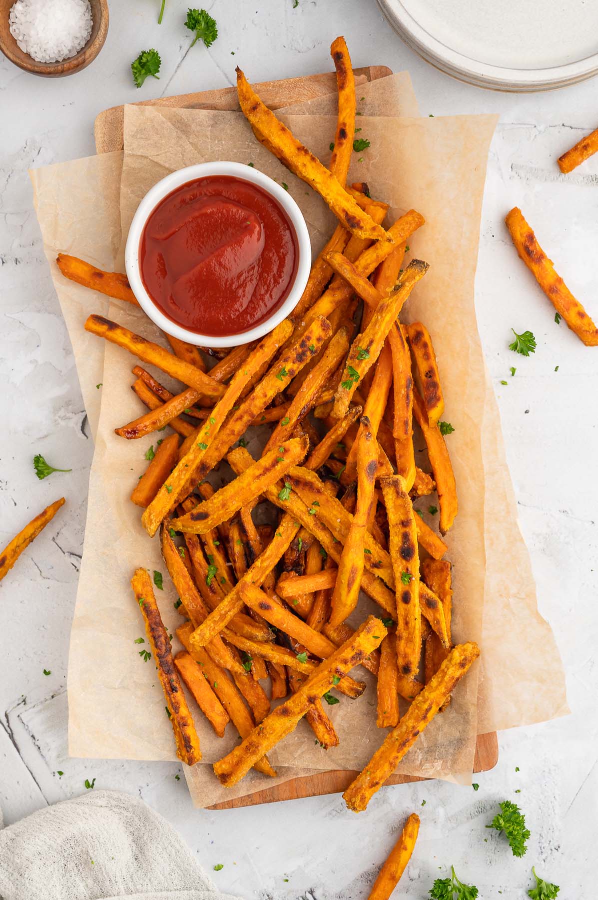 An overhead shot of baked sweet potato fries on a parchment lined platter with a white bowl of ketchup on the platter on a grey background