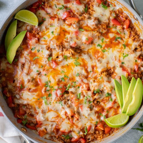 An overhead shot of a taco skillet in a large pan garnished with avocado slices and lime wedges on a grey background