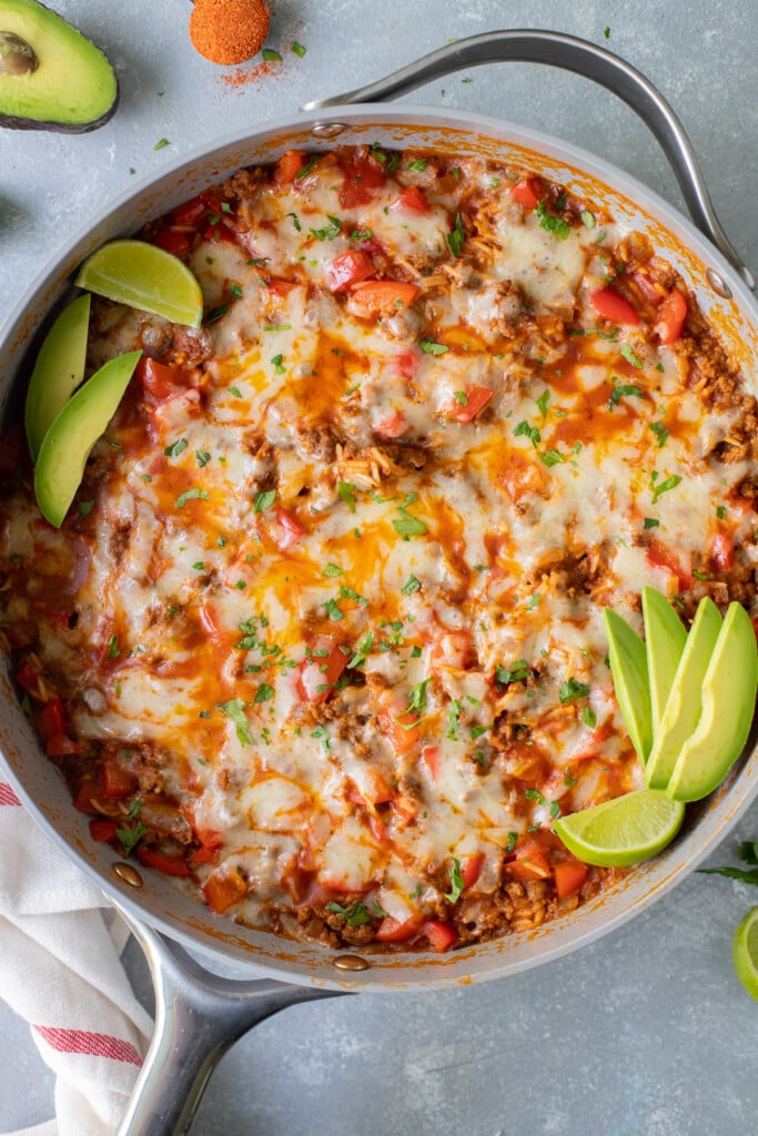An overhead shot of a taco skillet in a large pan garnished with avocado slices and lime wedges on a grey background