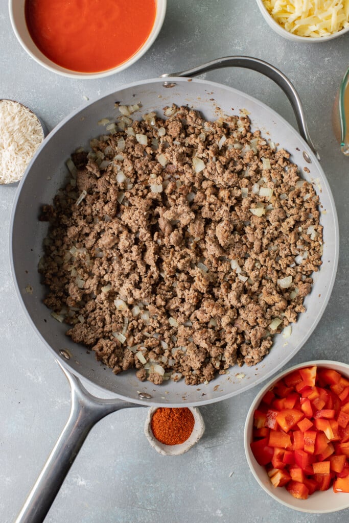 An overhead shot of browned ground beef, onion, and garlic in a skillet with remaining ingredients for taco skillet on the side