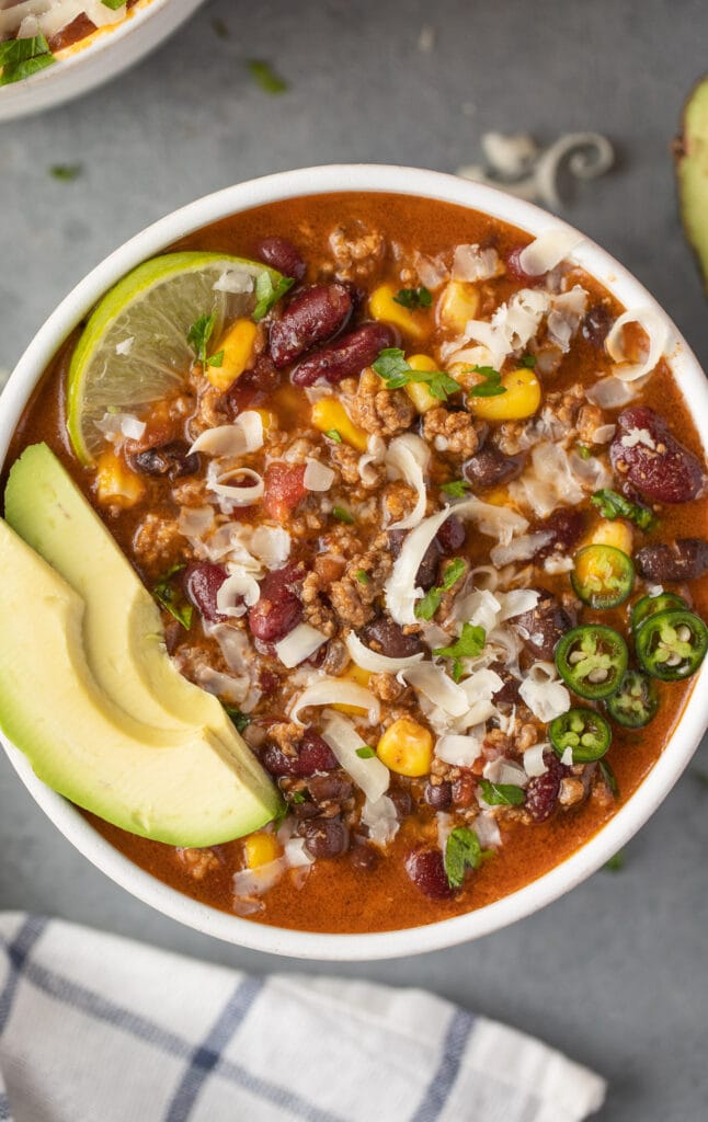 A close up bowl of taco soup with avocado and lime in the bowl on a grey background