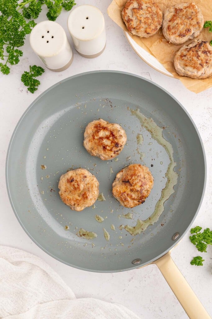 Three Turkey Breakfast Sausage patties in a pan with more patties on a plate on the side.