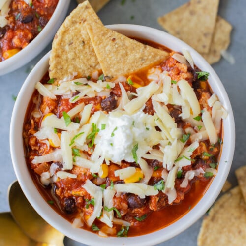 An overhead shot of a bowl of ground turkey chili with white cheese on top and two tortilla chips dipped in with gold spoons and more chips on the side