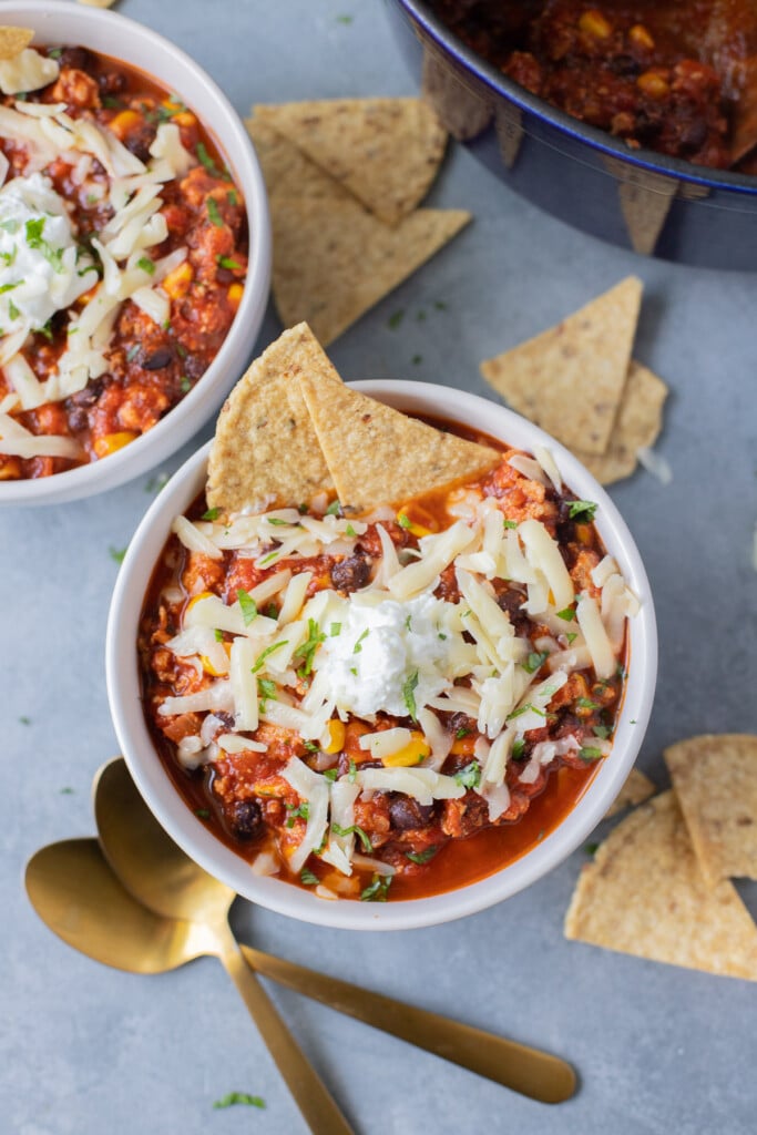 An overhead shot of a bowl of ground turkey chili with white cheese on top and two tortilla chips dipped in with another bowl of chili, gold spoons, and more chips on the side