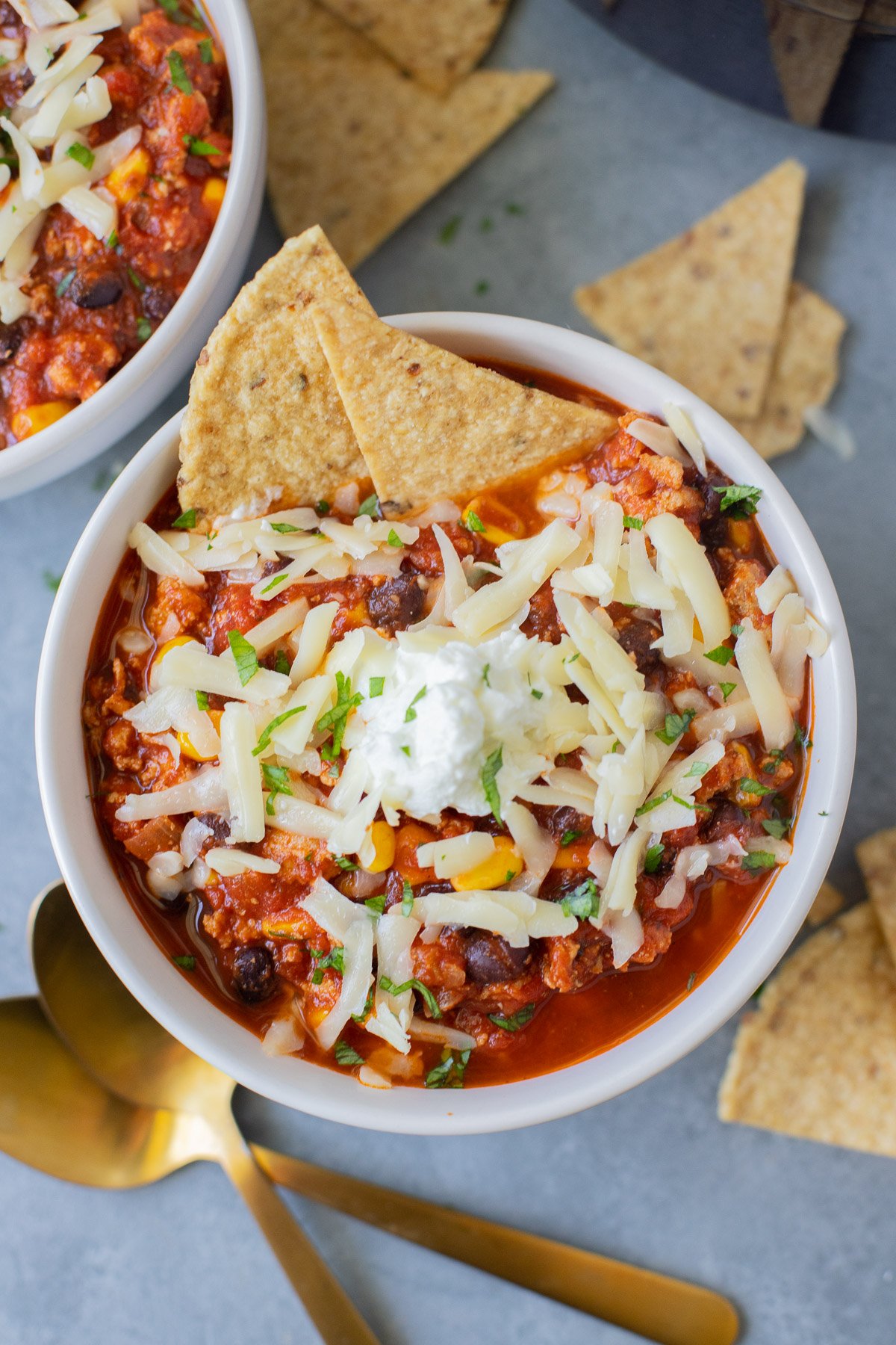An overhead shot of a bowl of ground turkey chili with white cheese on top and two tortilla chips dipped in with gold spoons and more chips on the side