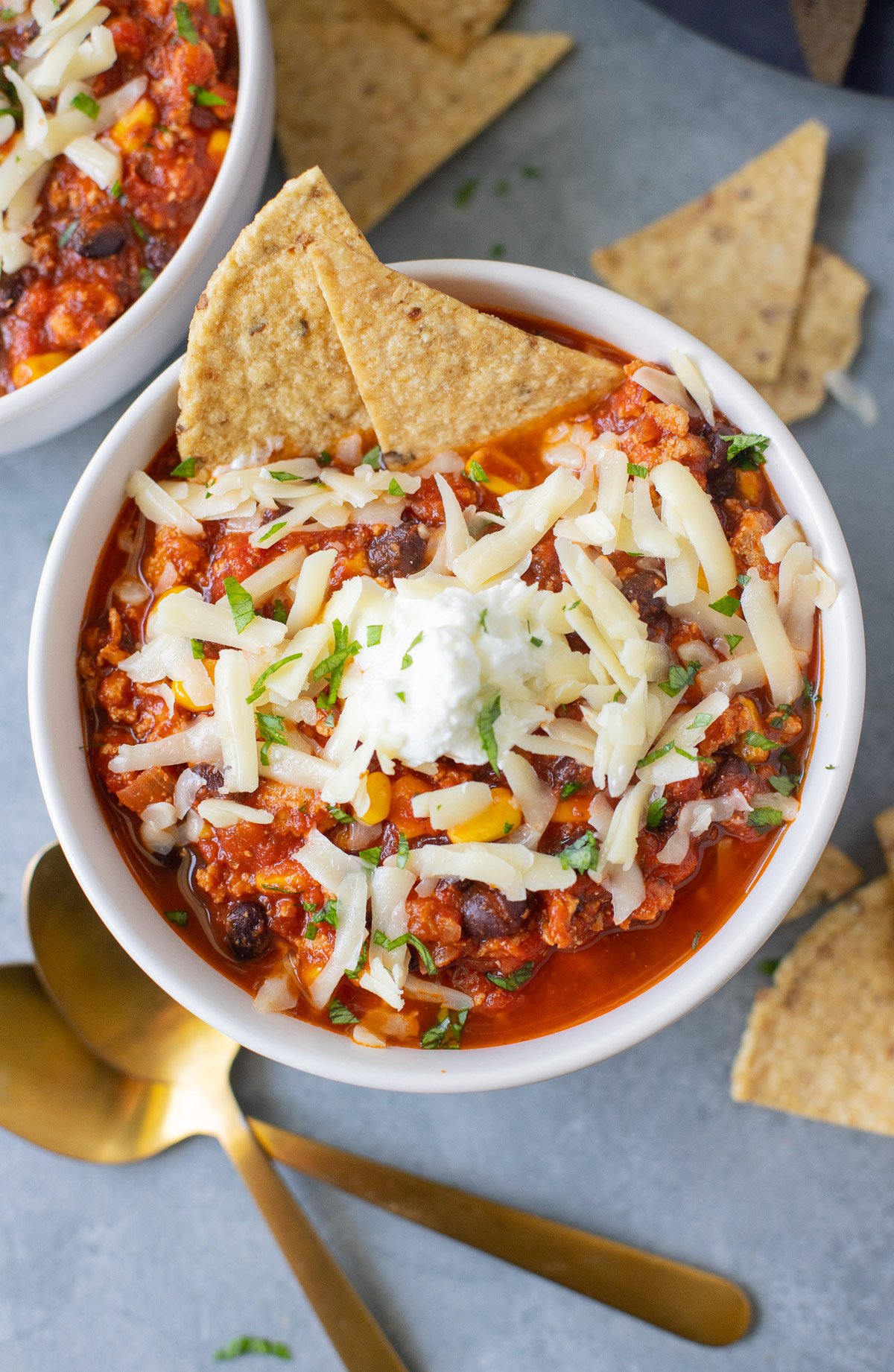 A bowl of ground turkey chili with white cheese on top and two tortilla chips dipped in with gold spoons and more chips on the side
