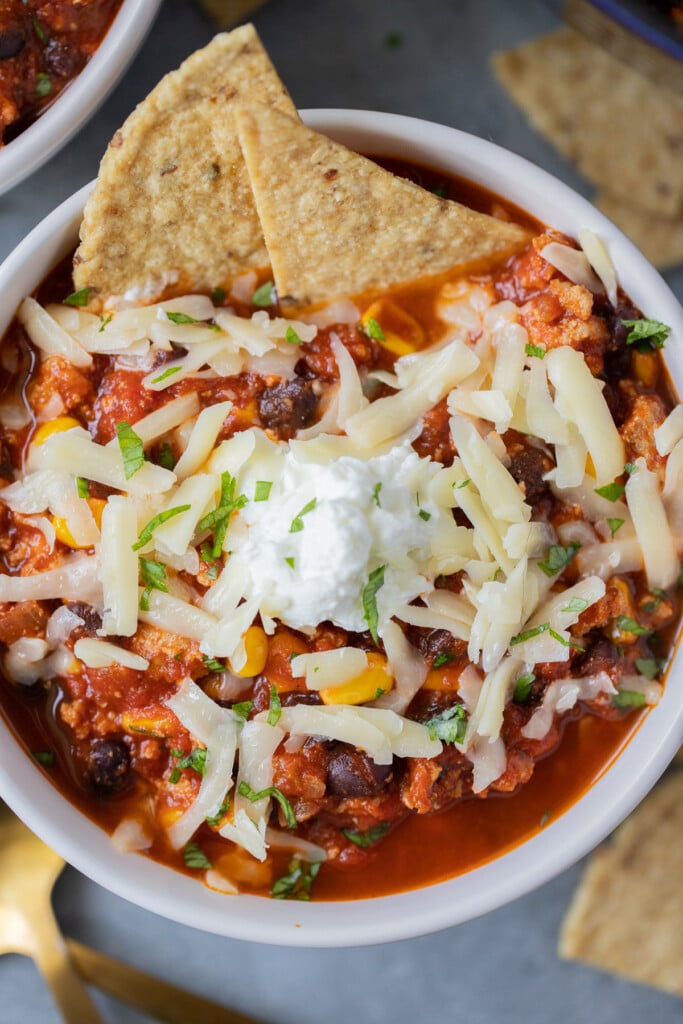 A close up of a bowl of ground turkey chili with white cheese on top and two tortilla chips dipped in