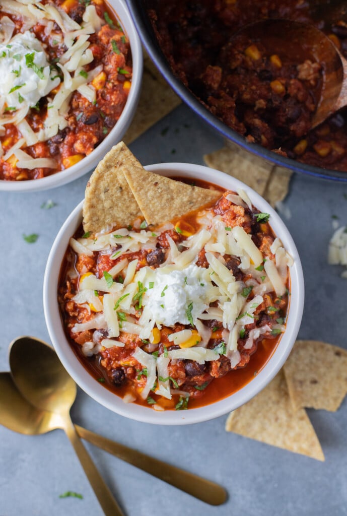 An overhead shot of a bowl of ground turkey chili with white cheese on top and two tortilla chips dipped in with another bowl and pot of chili, gold spoons, and more chips on the side