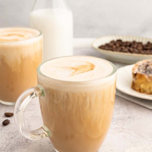A vanilla latte in a clear mug with another mug, bottle of milk and cinnamon roll in the background.
