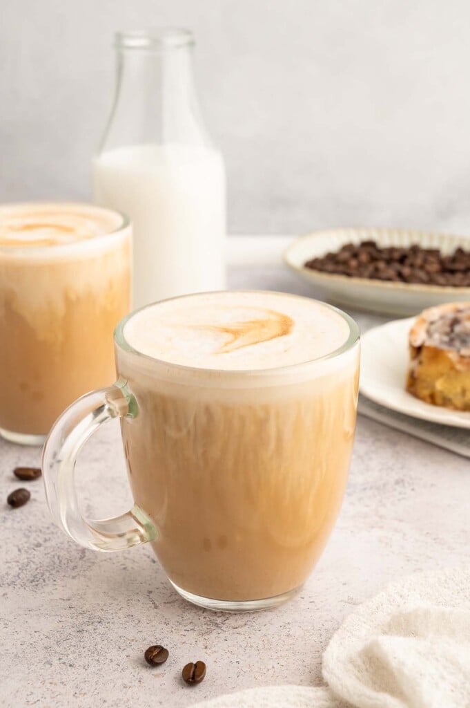 A vanilla latte in a clear mug with another mug, bottle of milk and cinnamon roll in the background.