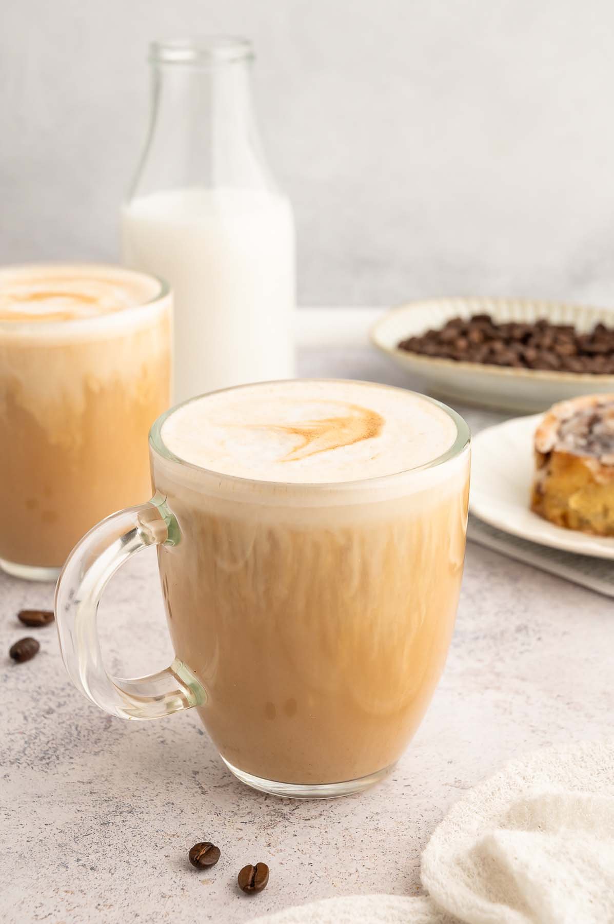 A vanilla latte in a clear mug with another mug, bottle of milk and cinnamon roll in the background.