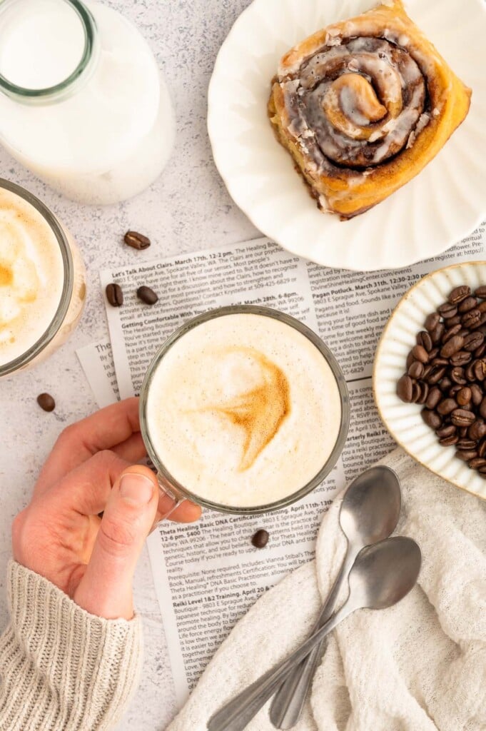 A hand holding a skinny vanilla latte in a mug with a cinnamon roll on the side. 