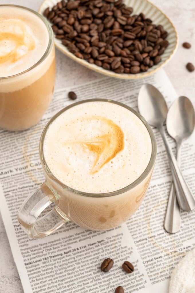 Two clear mugs with healthy vanilla lattes with spoons and a bowl of espresso beans on the side.