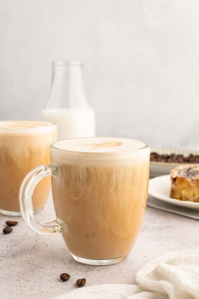 A clear mug filled with a healthy vanilla latte with another mug and cinnamon roll in the background. 