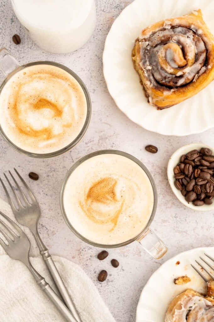 An overhead shot of two healthy vanilla lattes with a cinnamon roll on the side.