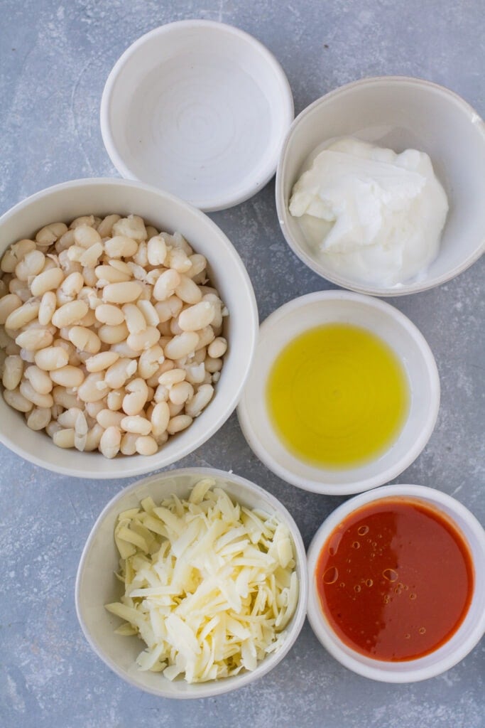 Ingredients for Vegetarian Buffalo Dip in small white bowls on a grey background: water, nonfat greek yogurt, extra virgin olive oil, buffalo sauce, cheddar cheese, cooked cannellini beans