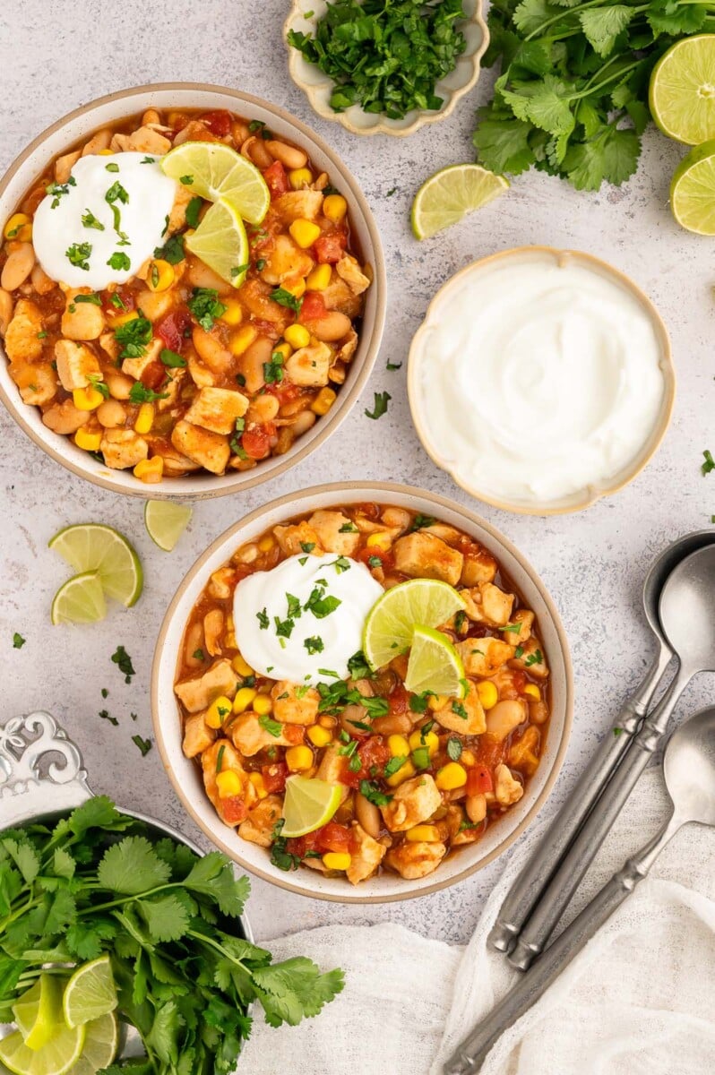Two bowls of white bean chicken chili in white bowls on a grey background with fresh cilantro and lime wedges on the side.