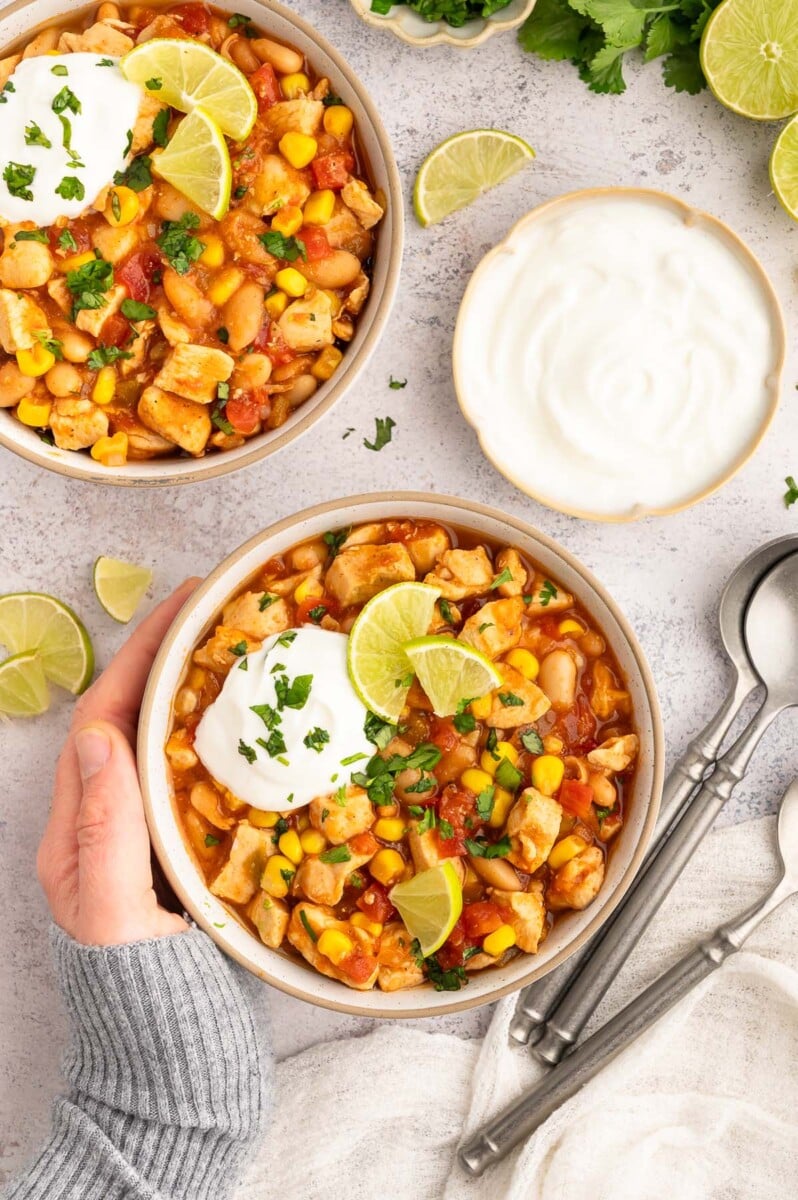 Two bowls of white bean chicken chili in white bowls on a grey background with a hand cupping one of the bowls.