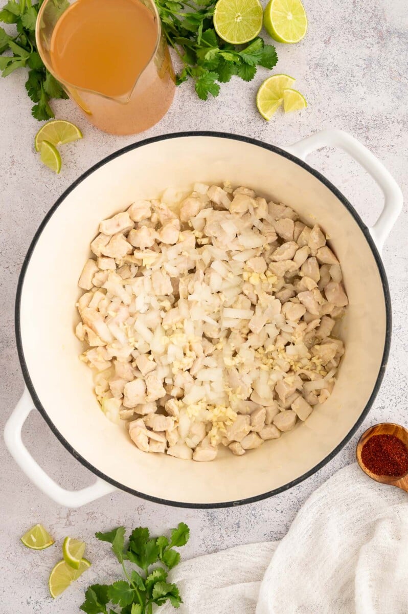 A dutch oven with cubed cooked chicken, garlic, and diced onion in it on a grey background.