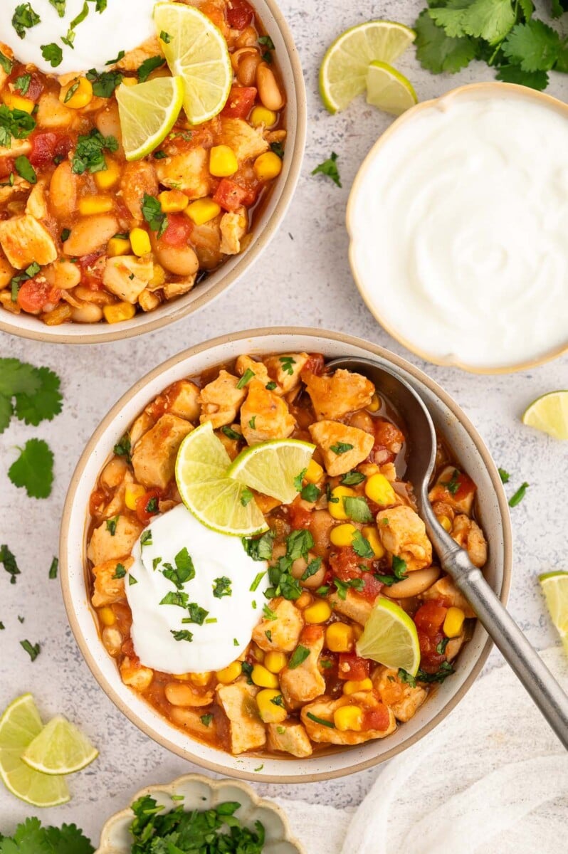 A close up bowl of white bean chicken chili with a spoon in it on a grey background with another bowl of chili on the side.