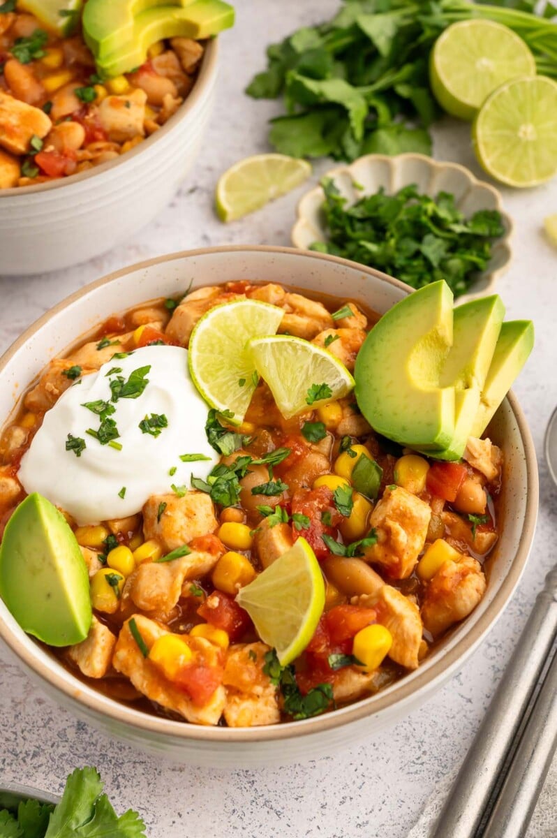 A close up bowl of white bean chicken chili in a white bowl on a grey background with a bowl of chili, limes, and fresh cilantro on the side.