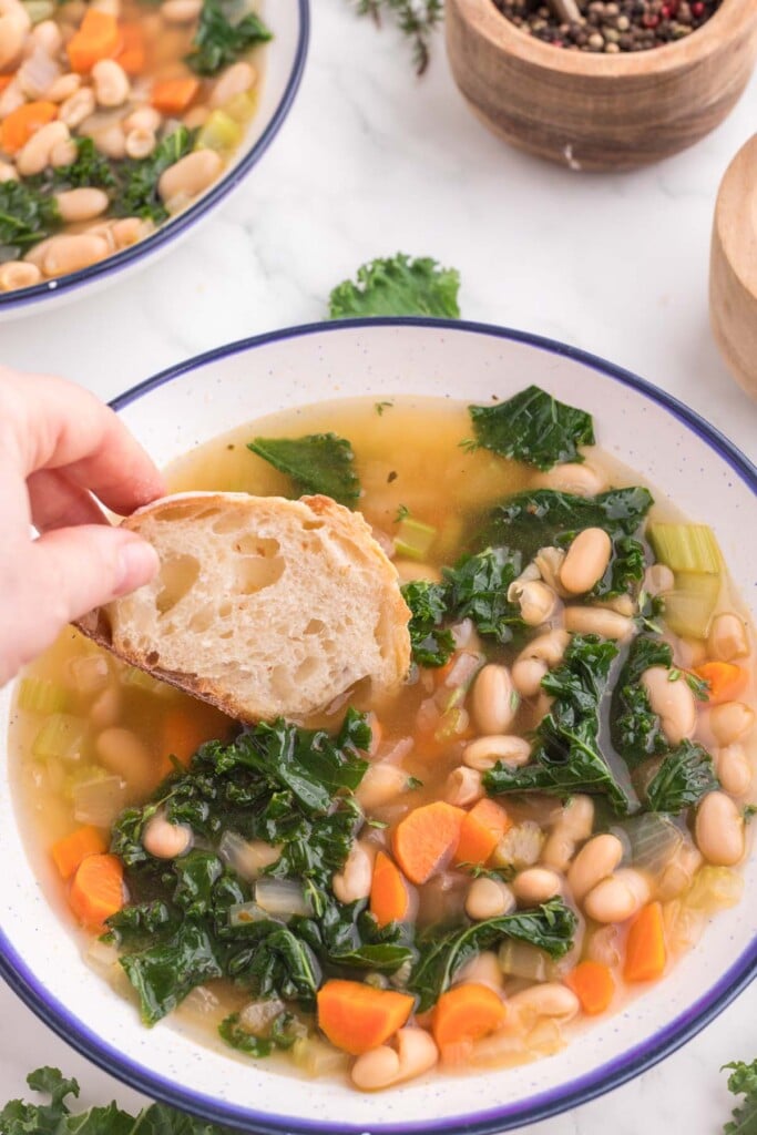 A hand about to dip a piece of bread into a bowl of Tuscan white bean soup on a white counter.
