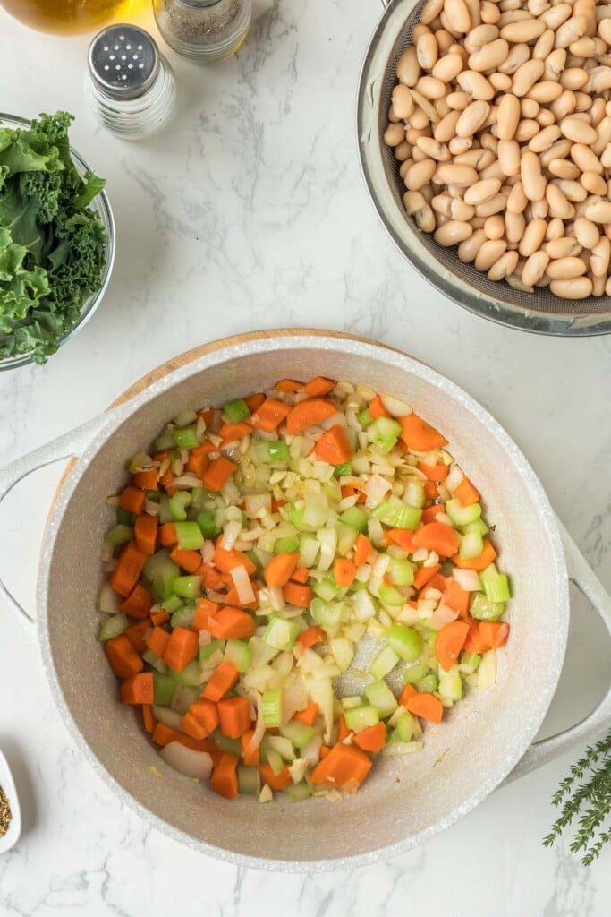 Onion, Garlic, Carrots, and Celery in a dutch oven on a white counter with more ingredients for tuscan white bean soup on the side. 