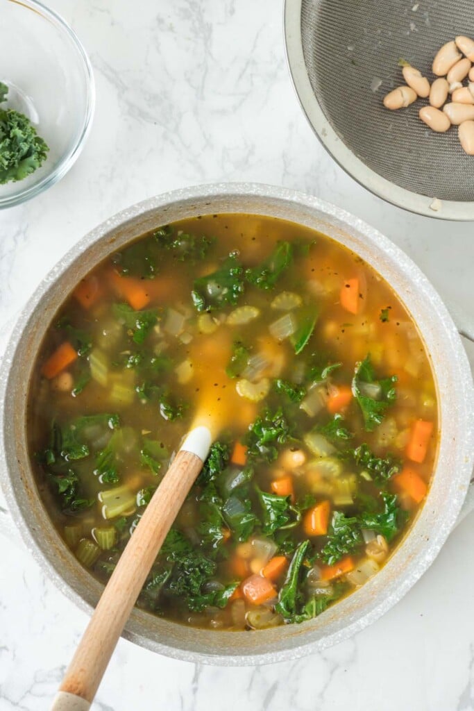 Tuscan white bean soup in a dutch oven being stirred with a wooden spoon on a white counter.