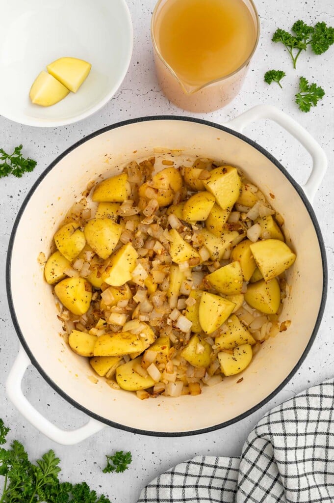 Ground Italian sausage, onion, garlic, potatoes, and spices in a dutch oven on a white background with a cup of broth on the side to add and make zuppa toscana
