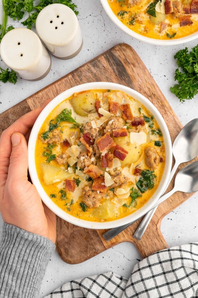 Healthy Zuppa Toscana Soup in a white bowl with a hand cupping the side on a wooden cutting board on a white counter