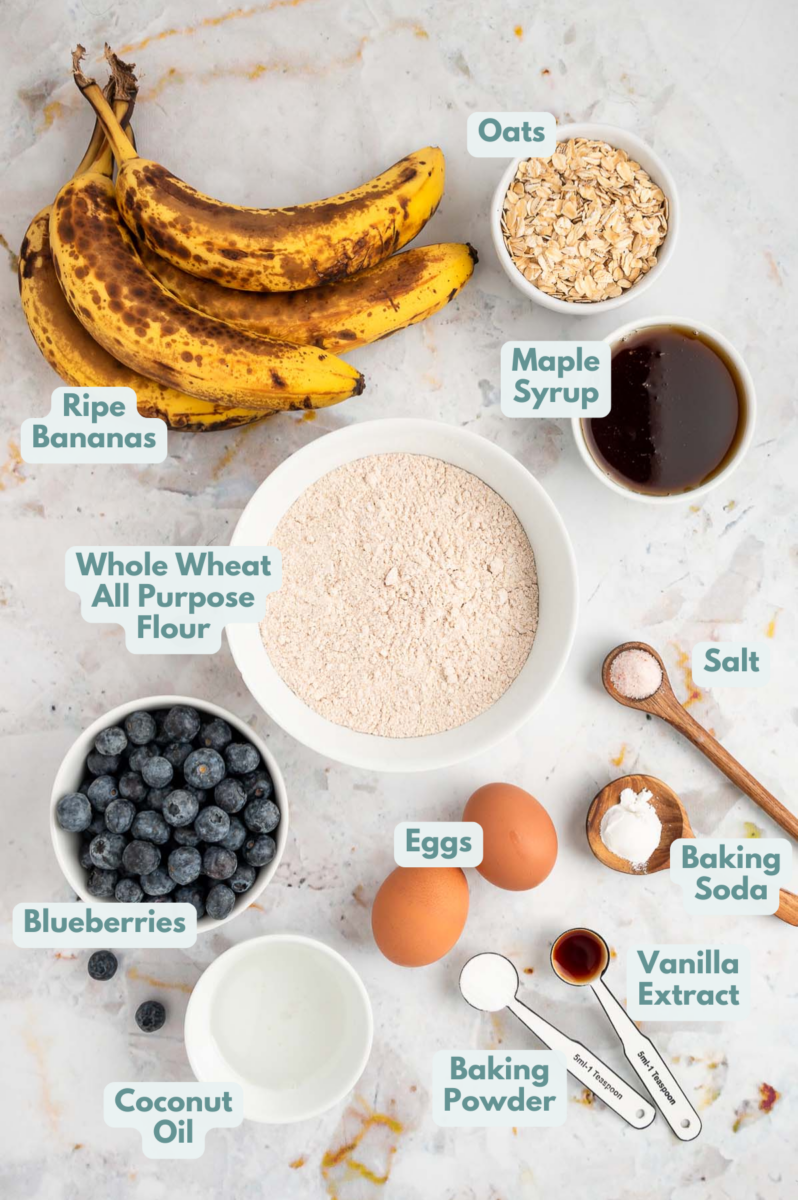 Ingredients for blueberry banana muffins in white bowls: old-fashioned oats, whole wheat all-purpose flour, baking soda, baking powder, sea salt, ripe bananas, eggs, maple syrup, coconut oil, vanilla extract, fresh blueberries
