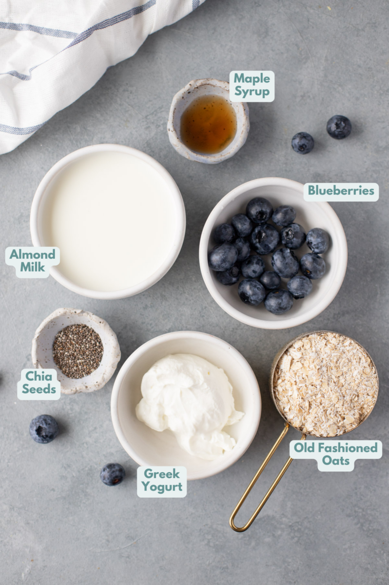 Ingredients for blueberry overnight oats in small white bowls on a grey background: old fashioned rolled oats, greek yogurt, almond milk, blueberries, maple syrup, chia seeds (optional)