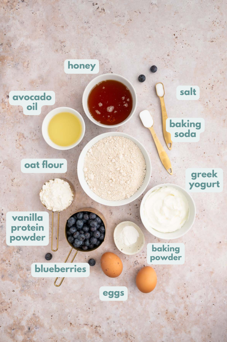 Ingredients for Blueberry Protein Muffins in small bowls: greek yogurt, honey, eggs, avocado oil, oat flour, vanilla whey protein powder, baking powder, baking soda, salt, blueberries