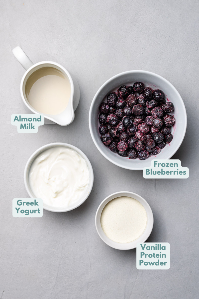 Ingredients for the Best Blueberry Smoothie Recipe on a light grey background: almond milk, frozen blueberries, greek yogurt, vanilla protein powder.