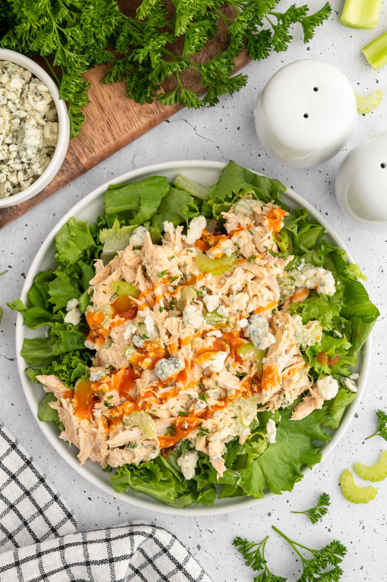 High Protein Buffalo chicken salad with blue cheese crumbles on a bed of lettuce in a white bowl on a grey background.