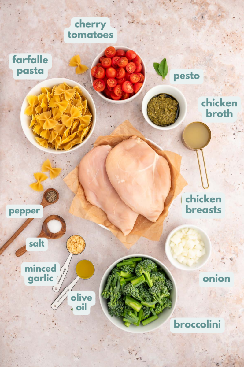 Ingredients for Chicken Pesto Pasta in small bowls: olive oil, chicken breasts, salt, pepper, chicken broth, farfalle pasta, minced garlic, minced onion, cherry tomatoes, broccolini, basil pesto