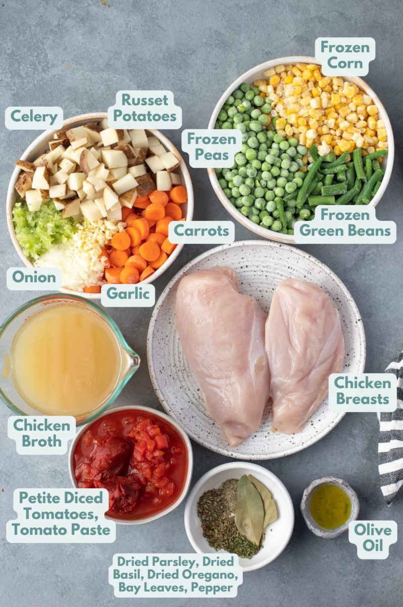 Ingredients for chicken and vegetable soup in white bowls on a blue background: olive oil, garlic, onions, celery, carrots, russet potatoes, chicken breasts, chicken broth, canned petite diced tomatoes, tomato paste, bay leaves, dried parsley, dried basil, dried oregano, frozen corn, frozen peas, frozen green beans