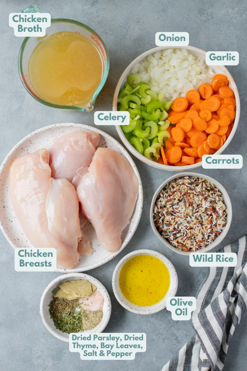 Ingredients for chicken and wild rice soup in white bowls: olive oil, celery, onion, carrots, garlic, salt & pepper, dried parsley, dried thyme, bay leaves, chicken breast, chicken broth, wild rice blend