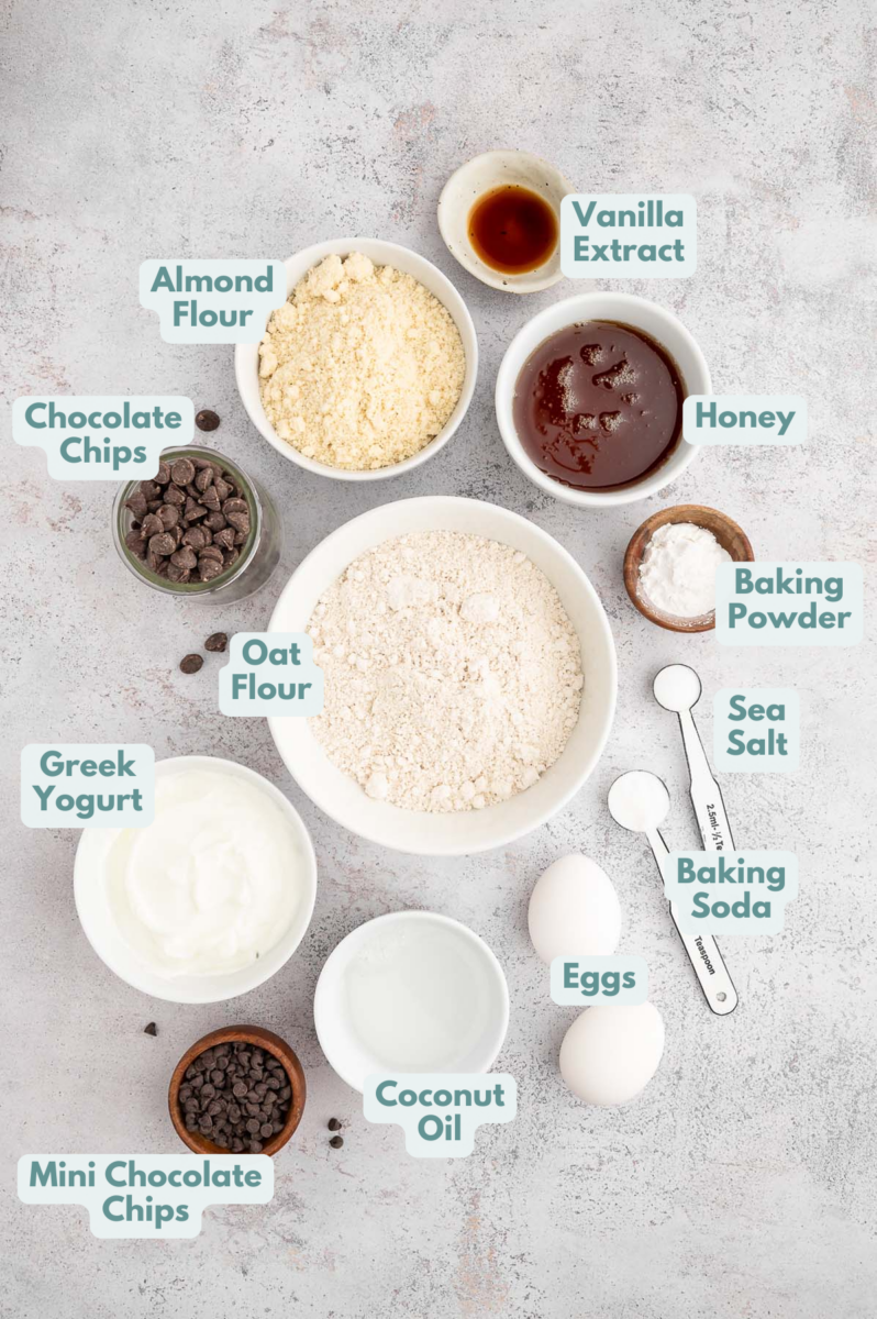 Ingredients for healthy chocolate chip muffins in white bowls: oat flour, almond flour, baking powder, baking soda, sea salt, nonfat greek yogurt, honey, eggs, coconut oil, vanilla extract, chocolate chips, mini chocolate chips