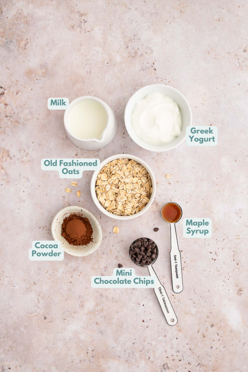 Ingredients for chocolate overnight oats in small bowls: old fashioned rolled oats, nonfat greek yogurt, milk, cocoa powder, mini chocolate chips, maple syrup