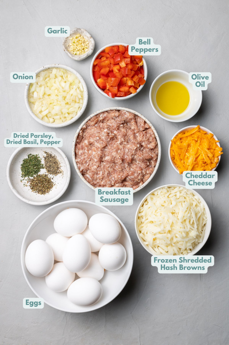 Ingredients for breakfast hash brown casserole in white bowls: olive oil, breakfast sausage, garlic, onions, bell pepper, eggs, dried parsley, dried basil, pepper, frozen shredded hash browns, cheddar cheese