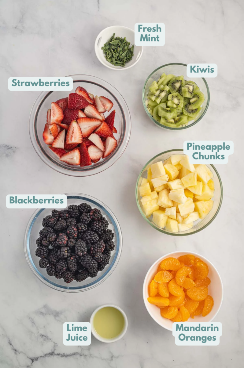 Ingredients for Fruit Salad in small bowls: strawberries, mandarin oranges, blackberries, kiwis, pineapple, lime juice, fresh mint.