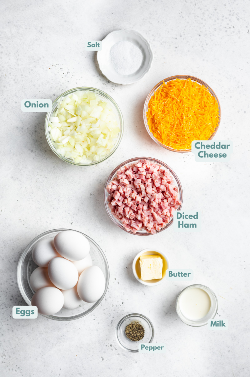Ingredients for ham and cheese frittata in white bowls: salted butter, yellow onion, eggs, whole milk, salt, pepper, shredded cheddar cheese, diced ham, fresh chives or fresh parsley