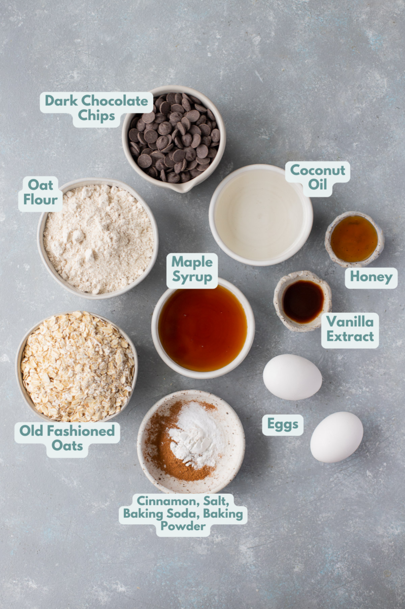 Ingredients for healthy oat flour cookies in small bowls on a grey counter: old fashioned rolled oats, oat flour, cinnamon, baking soda, baking powder, salt, coconut oil, maple syrup, honey, eggs, vanilla extract, dark chocolate chips