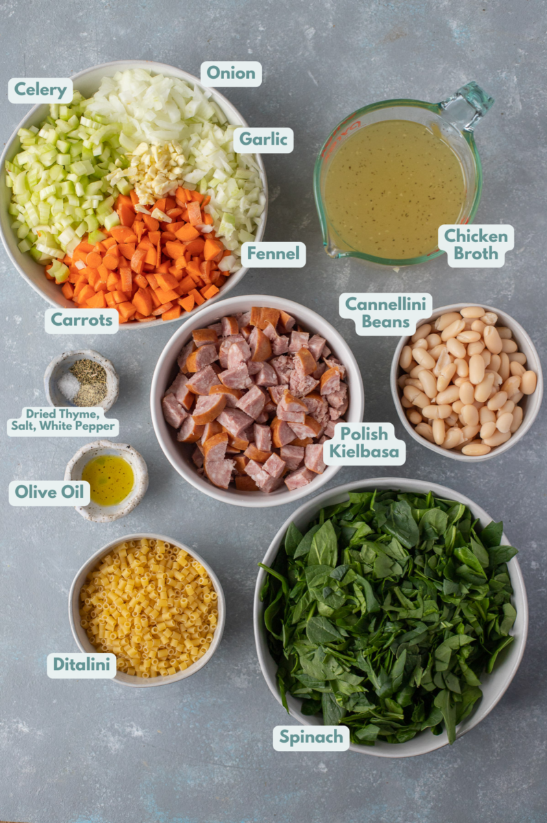 Ingredients for polish sausage soup in small bowls: olive oil, polish kielbasa, garlic, fennel, onion, carrots, celery, dried thyme, white pepper, cannellini beans, chicken broth, baby spinach, ditalini